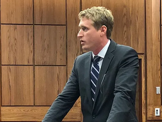 Man in dark suit and striped tie speaking in a wood-paneled room.