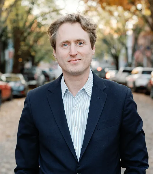 Portrait of a man in a dark blazer and striped shirt standing on a tree-lined street with parked cars.