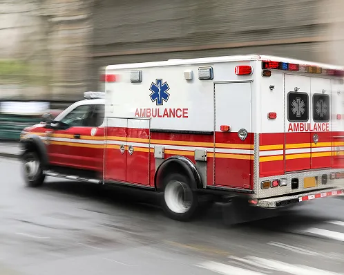 Red and white ambulance with emergency lights driving quickly on wet city street.