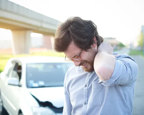 Man in glasses and light blue shirt holding neck in pain in front of damaged white car.