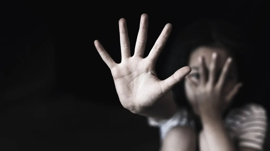 Child with outstretched hand in focus, face partially covered, dark background.