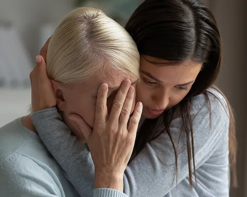 Two women embracing in a comforting manner, one visibly distressed with her face covered.