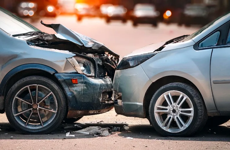 Two silver cars with front-end damage involved in a head-on collision on a street.