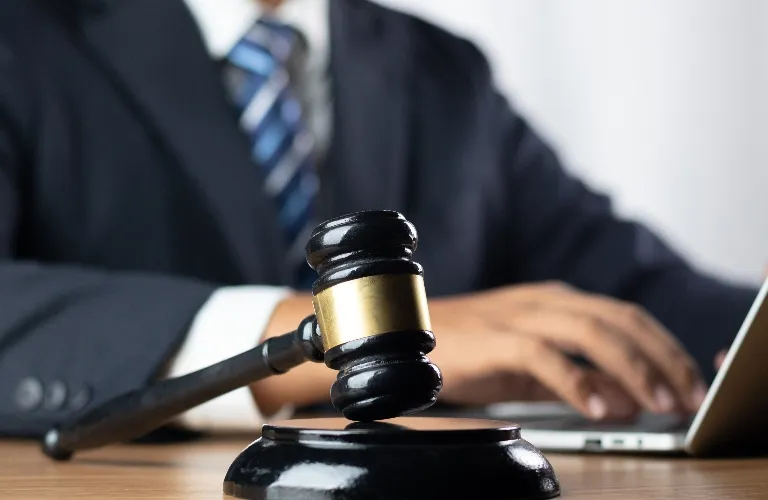 Close-up of a black judge's gavel with a gold band on a wooden desk, businessman typing on laptop in background.