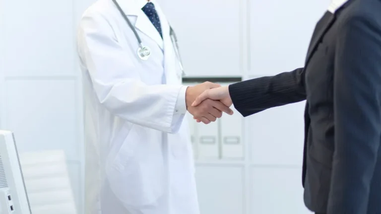 Doctor in white coat shaking hands with person in dark business suit.