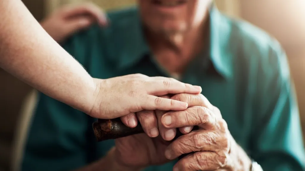 Close-up of a younger hand gently holding an elderly person's hand over a wooden cane.