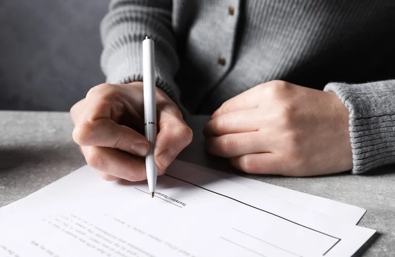 Person wearing a gray sweater holding a silver pen, signing a document on a gray surface.