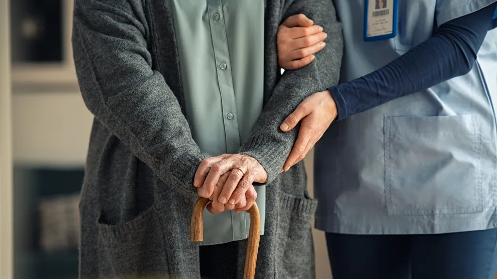Close-up of elderly person holding a cane, supported by a healthcare professional in blue scrubs.