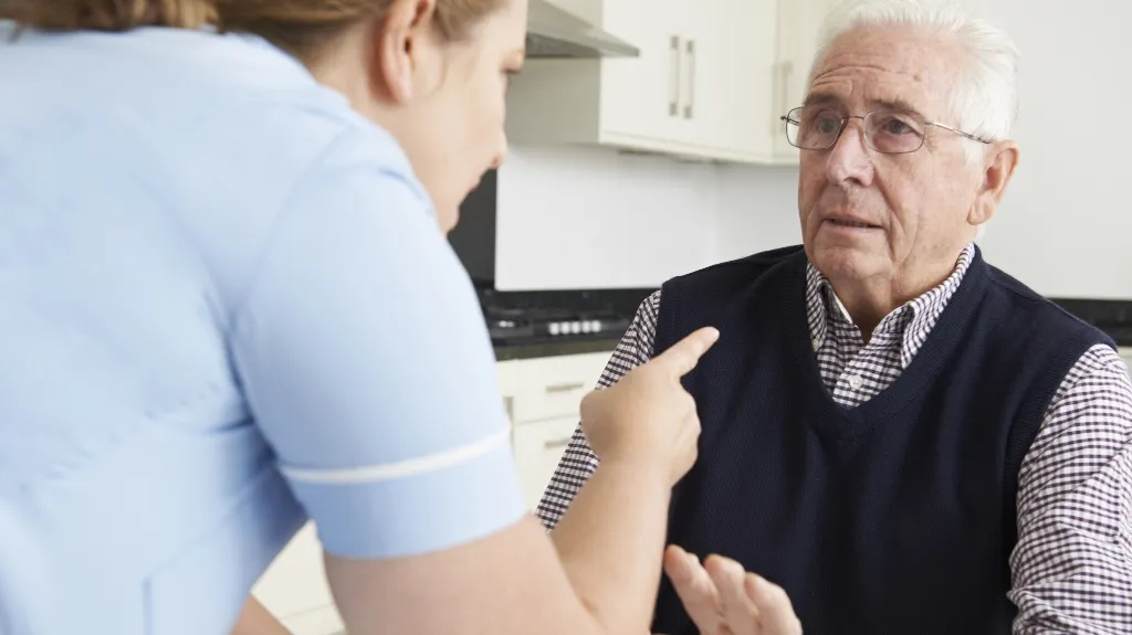 Healthcare professional in light blue uniform speaking to elderly man wearing glasses and a dark vest.
