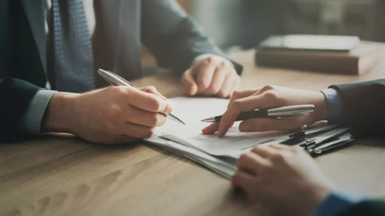Two professionals in suits reviewing and signing documents on a wooden table.